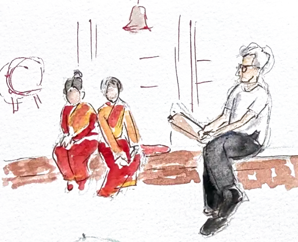 Two girls in red and gold sitting close together on a brick ledge, a man in a white shirt sitting apart from them reading, a temple bell hanging above